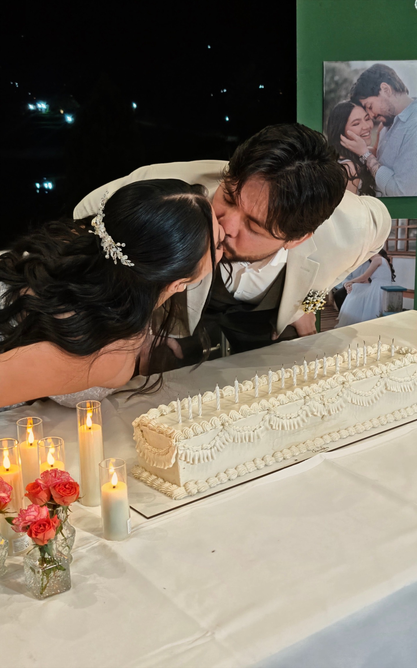 Beso de los novios sobre torta de bodas alargada en buttercream blanco con piping clásico y velas encendidas